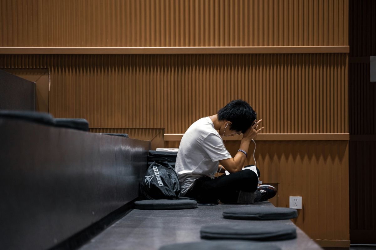 A person in a white shirt and black pants, wearing headphones, sits reading a book in the front row of an empty lecture hall.